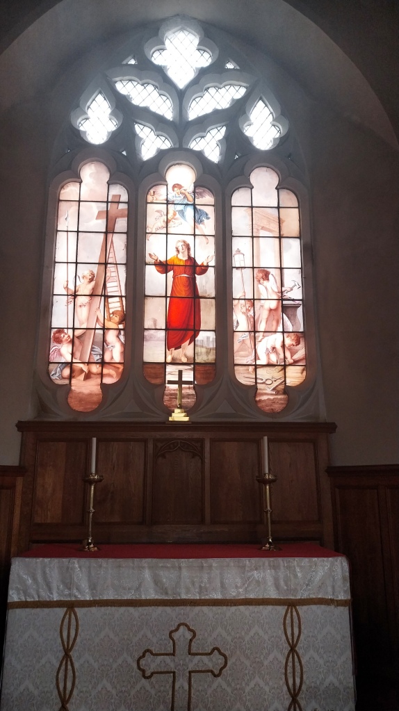 Altar and a triptych of glass windows. The central window shows a woman in a red dress. An angel dressed in blue flighs over her, as does a putto. The two other windows show putti at work. 